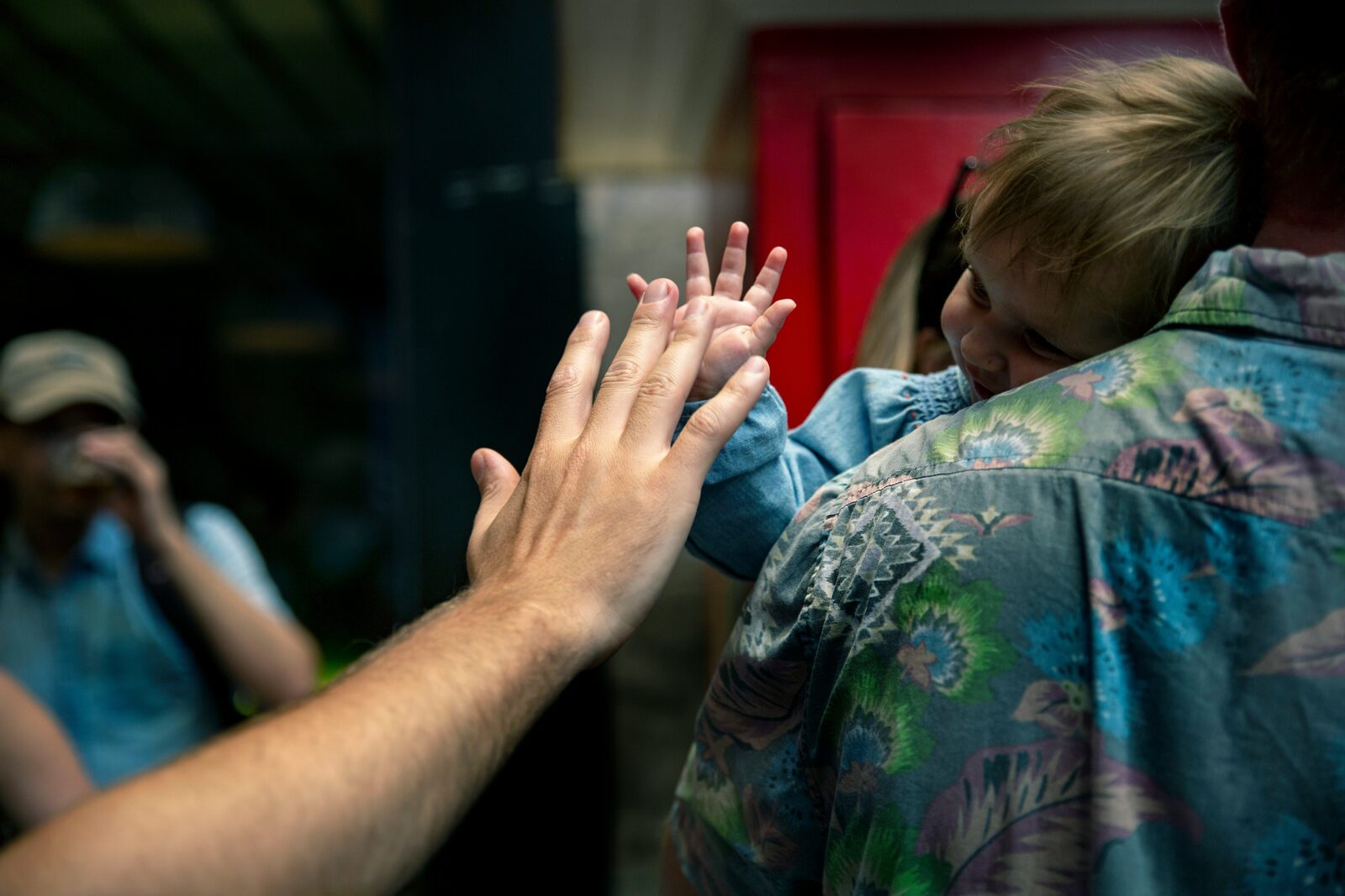 A small child held over someone's shoulder, pressing their palm against another person's outstretched hand